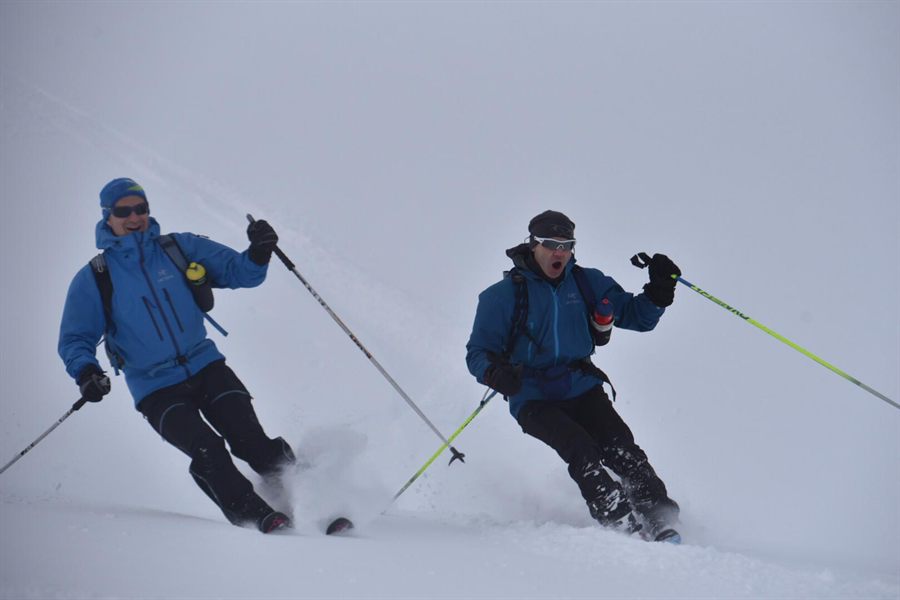 Two skiers in blue jackets making turns through soft snow in flat light