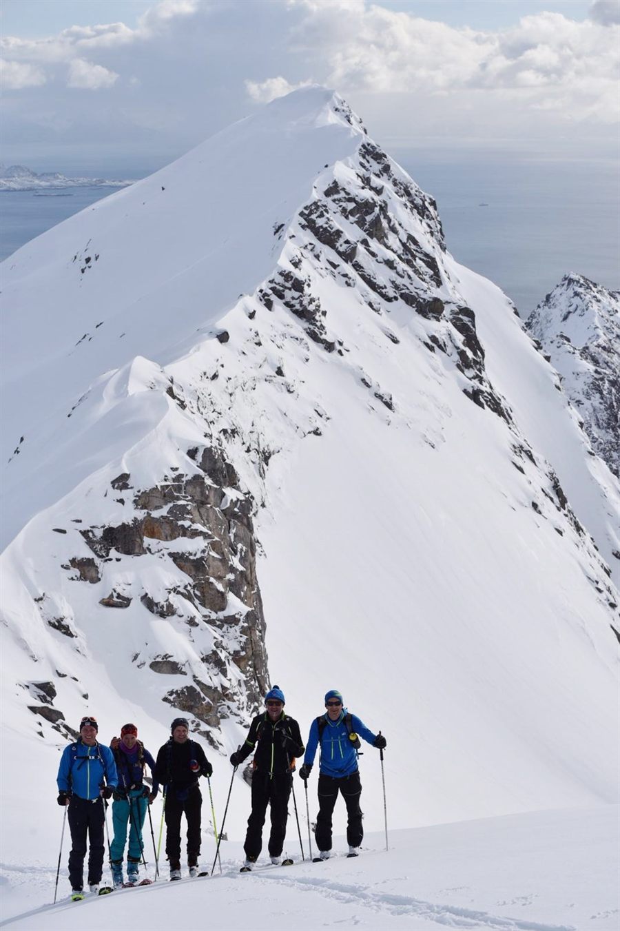 Five ski tourers standing on a ridge below a sharp snow covered peak, the sea behind them