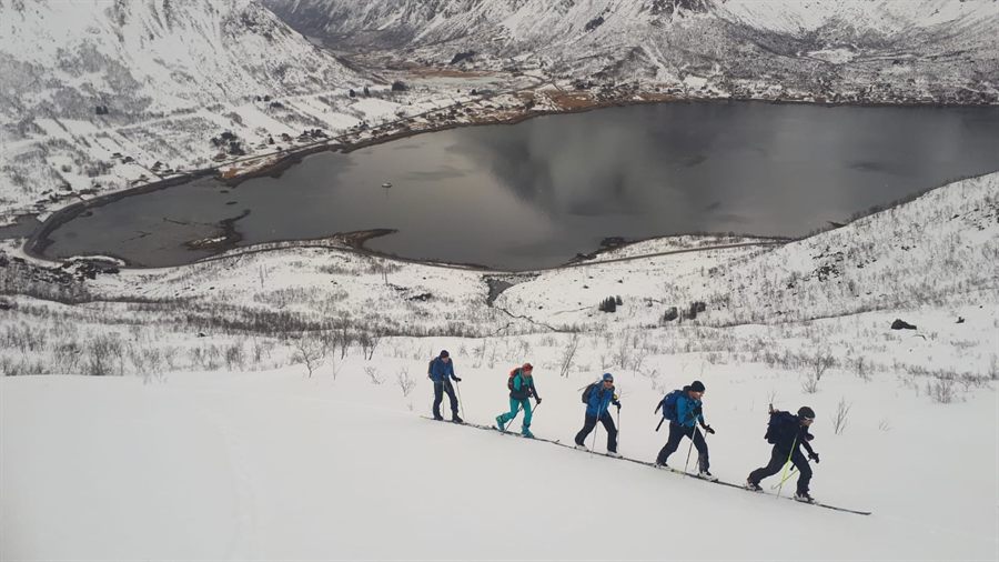 Five ski tourers traversing a snow slope high above a dark bay and a small village