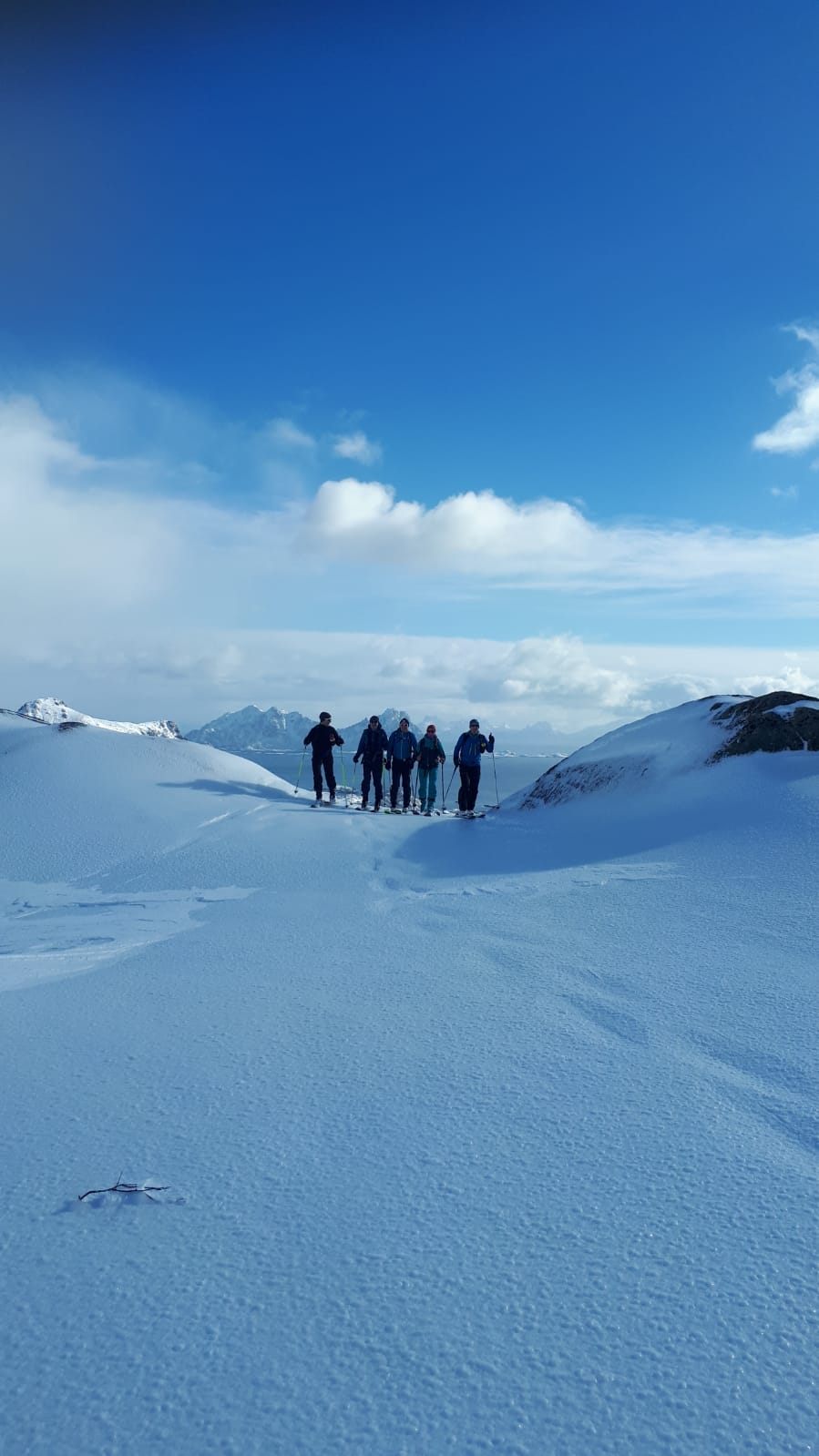 Five ski tourers on a broad snowy summit under a blue sky, distant peaks on the horizon
